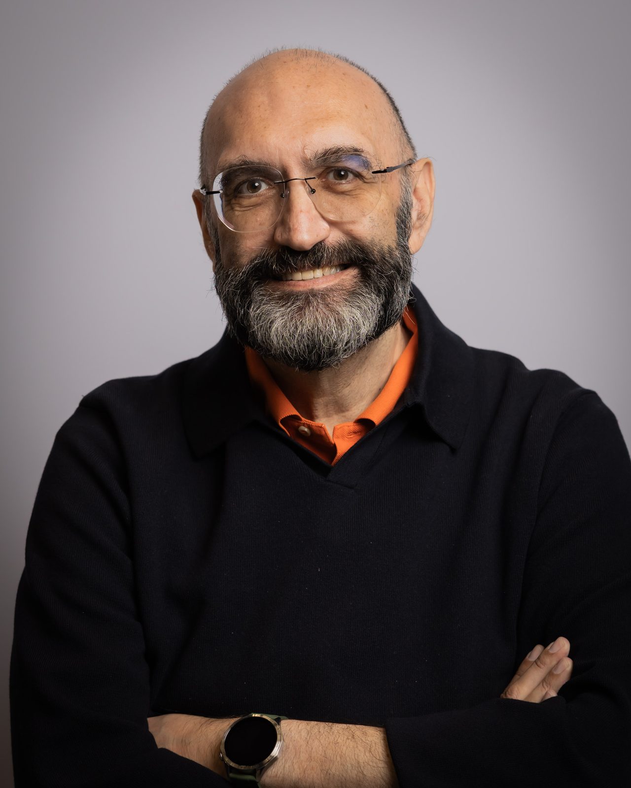 Malek Mroueh smiling in a dark jumper over an orange shirt, with his arms folded