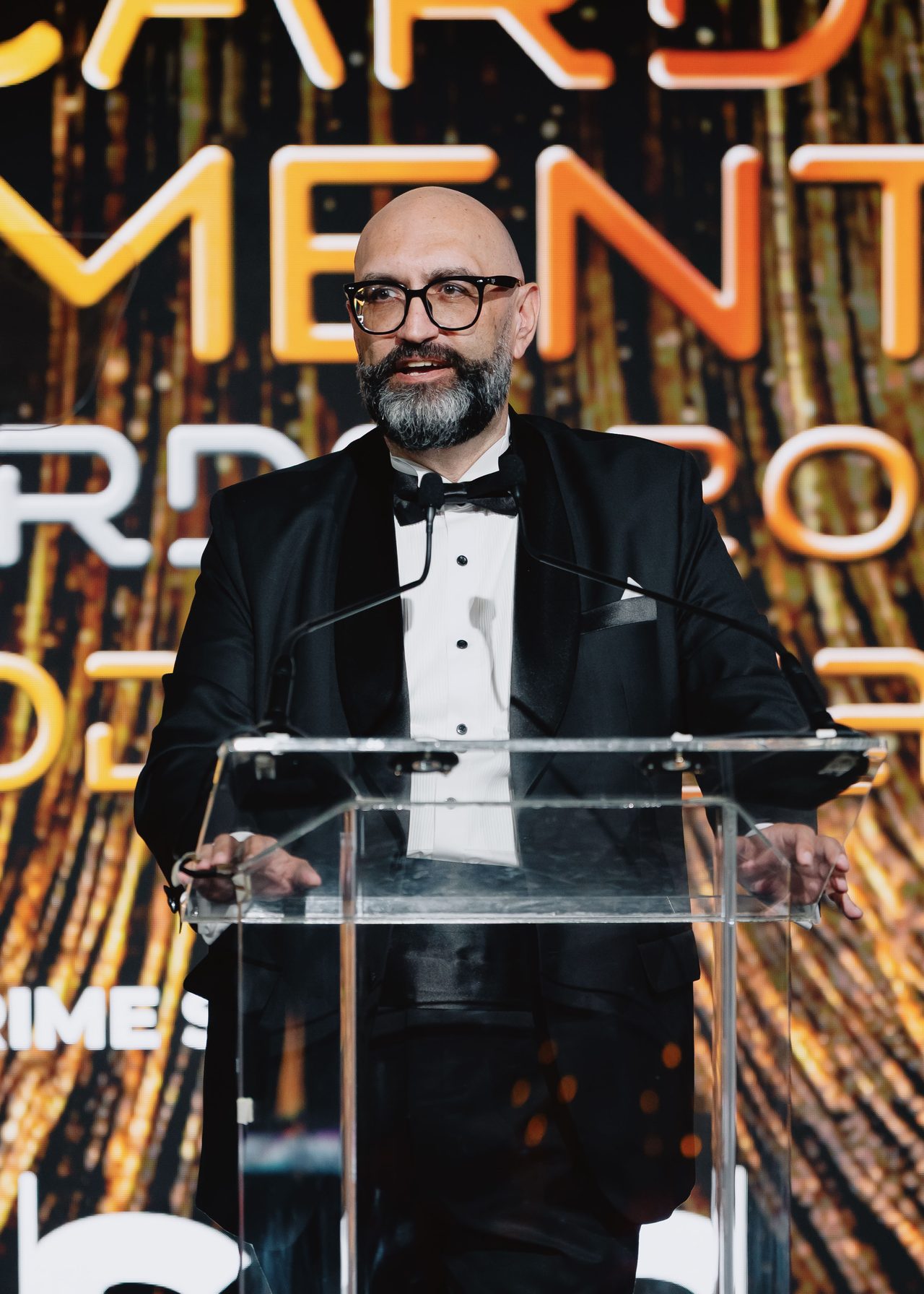 Malek Mroueh speaking at a clear lectern, wearing a tuxedo and bow tie